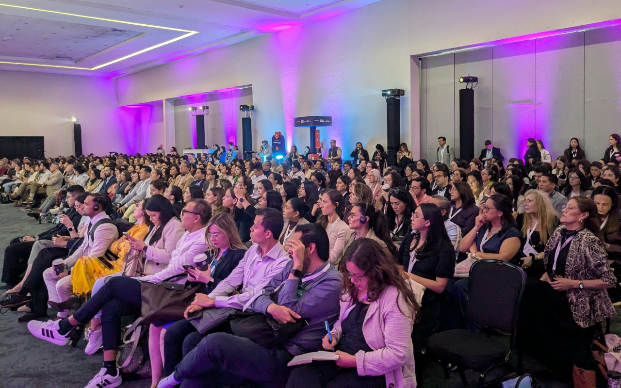 Large audience seated in a conference room with purple lighting, attentively watching a presentation or speaker at the front.