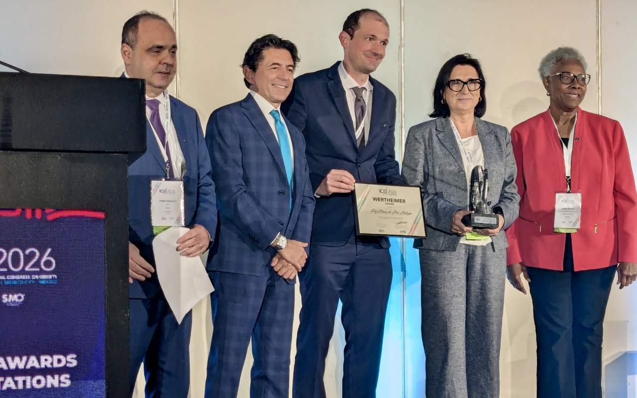 Five people stand onstage at an awards ceremony, with one man holding a certificate and one woman holding a trophy. All are dressed in business attire and facing the camera.