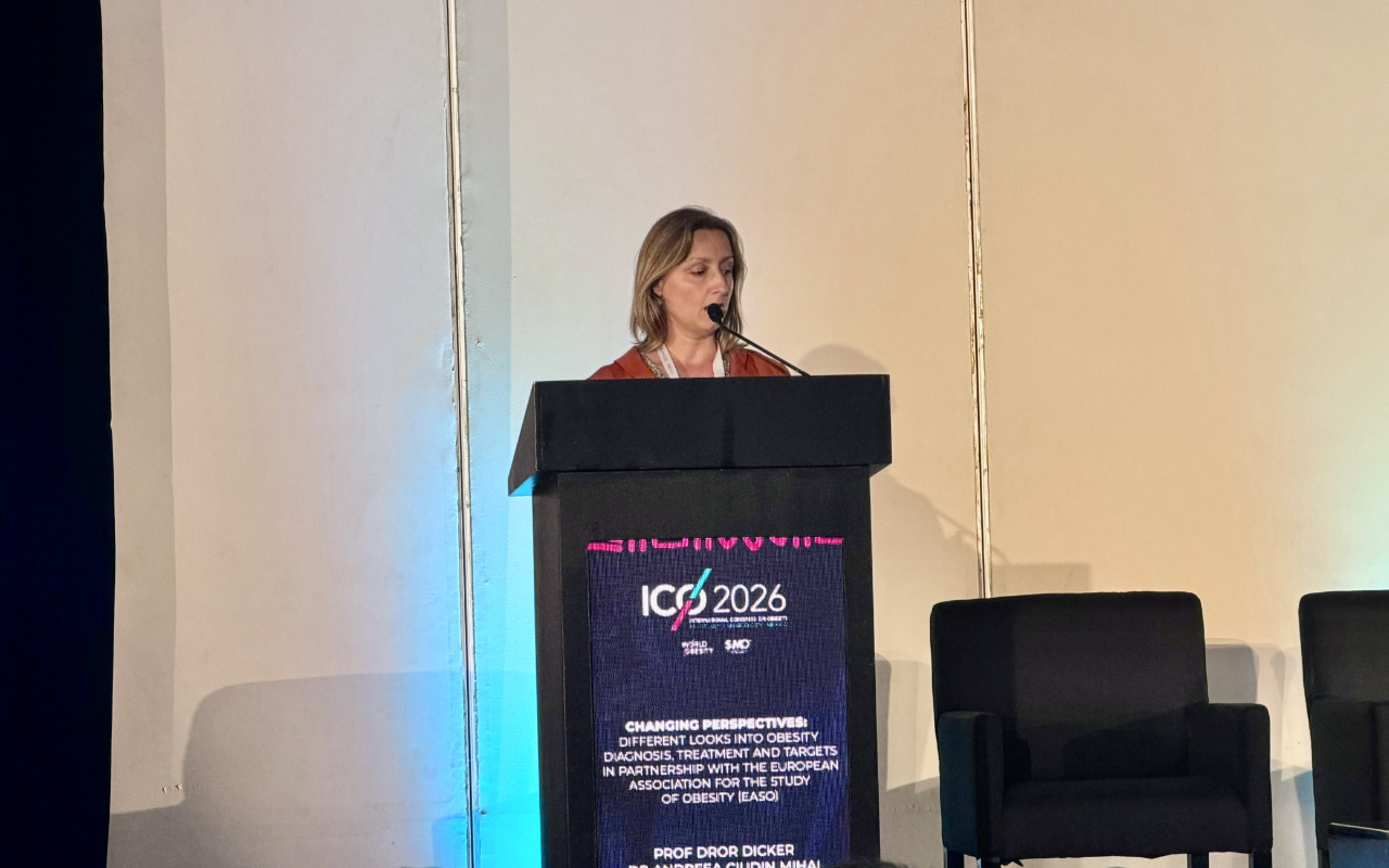A woman stands at a lectern giving a presentation at a conference. The lectern displays the ICO 2026 logo and event information. Two empty chairs are visible beside her.