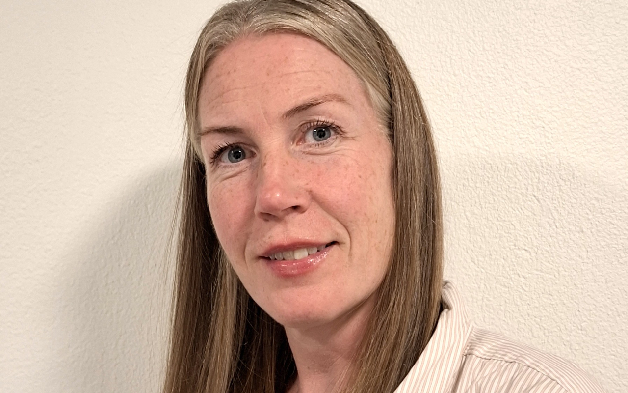 A woman with straight, light brown hair and a light complexion is standing in front of a white textured wall, looking at the camera and smiling slightly.