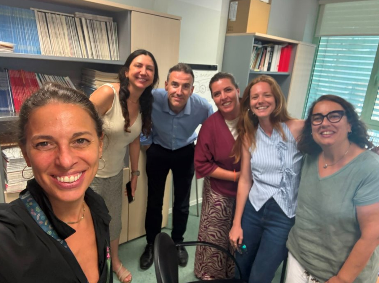 Six adults stand and smile together in an office with shelves of books and papers in the background.