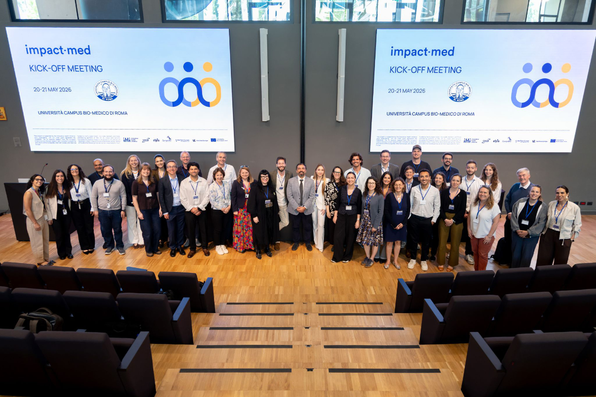 A large group of people pose for a photo in an auditorium during the “impact-med kick-off meeting”, with event details displayed on two screens behind them.