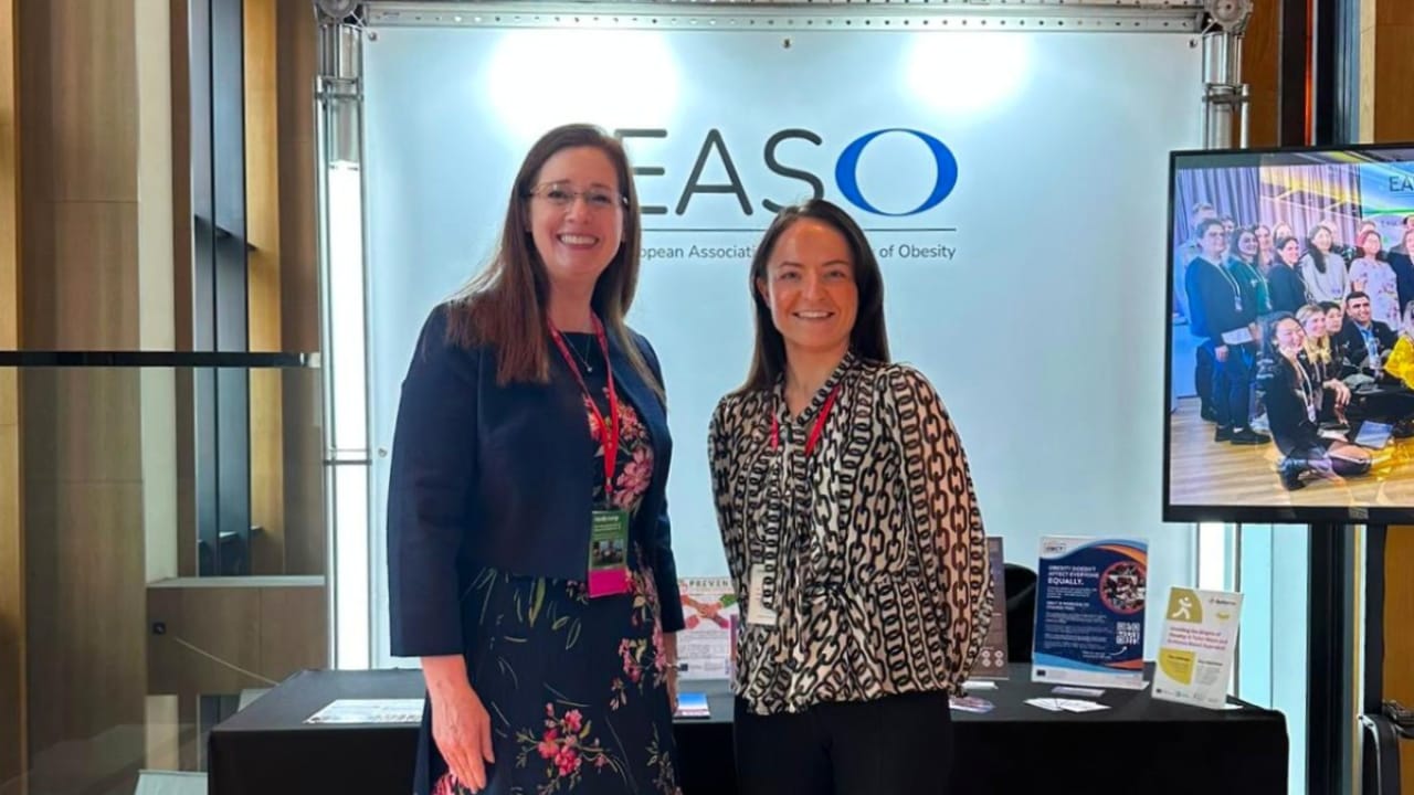 Two women stand smiling in front of an EASO stand display with informational materials on a table and a group photo on a screen.