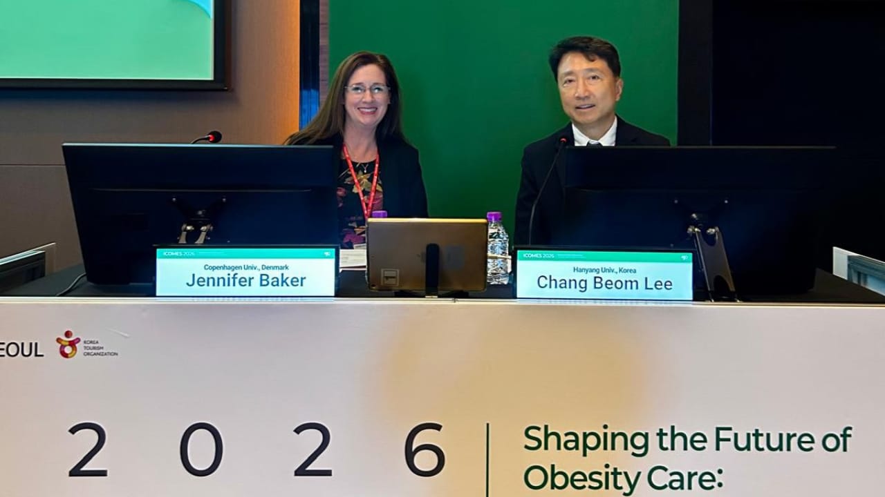 Two presenters, Jennifer Baker and Chang Beom Lee, sit at a conference table with nameplates during an event on the future of obesity care.