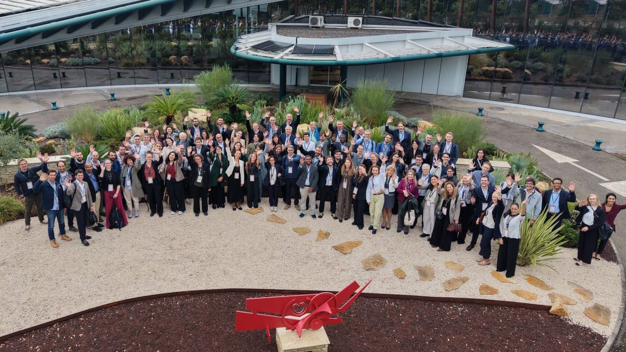 A large group of people standing in a courtyard outdoors and waving at the camera, with a modern building in the background and a red abstract sculpture in the foreground.